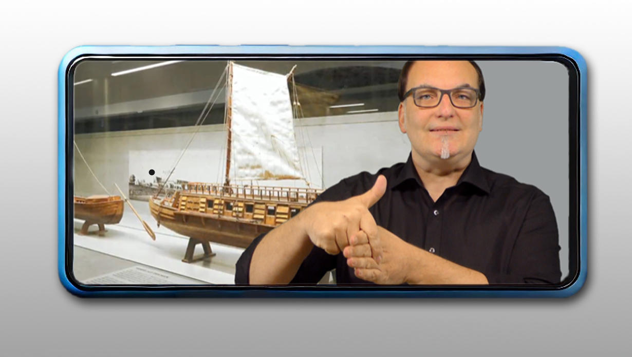 Rechts im Bild DGS-Dolmetscher, links im Hintergrund die Ausstellung in einem Museum, gezeigt im Display eines blauen Smartphones © Ingrid Glatz